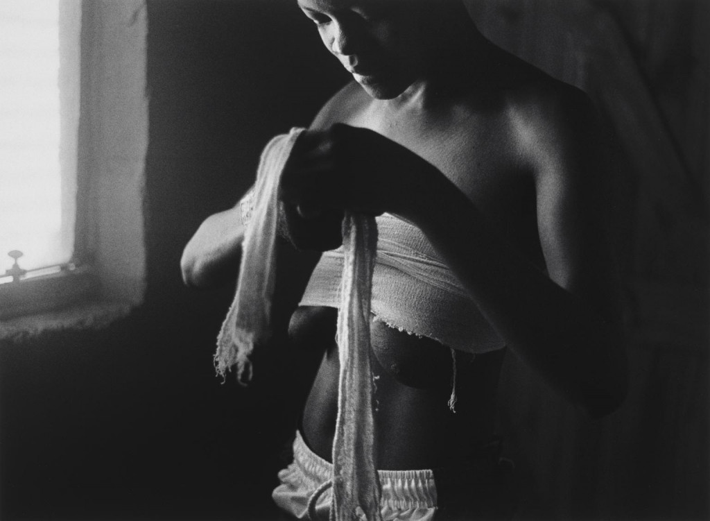 Zanele Muholi's black and white photograph, ID Crisis, shows a slim young woman standing in a gloomy room carefully wrapping bandages around her breasts in an attempt to disguise her anatomy, as light streams in from an adjacent window.